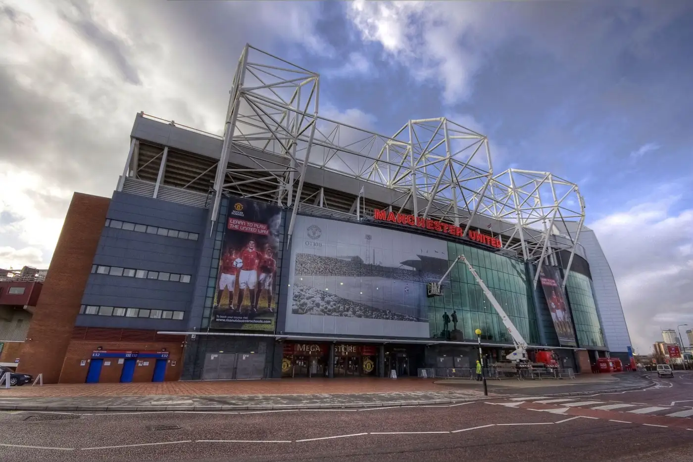 Façade vitrée d'Old Trafford surmontée des lettres rouges Manchester United et de la charpente blanche du toit, sous un ciel nuageux