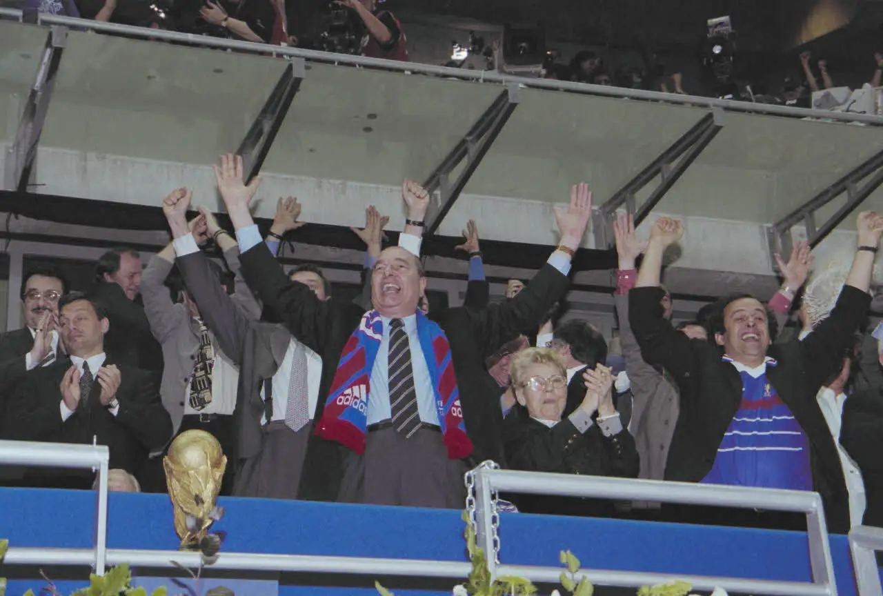 Jacques Chirac, une écharpe bleue et rouge autour du cou, et Michel Platini, en maillot de l'équipe de France, lèvent les bras dans la tribune présidentielle du Stade de France, le trophée de la Coupe du monde au premier plan