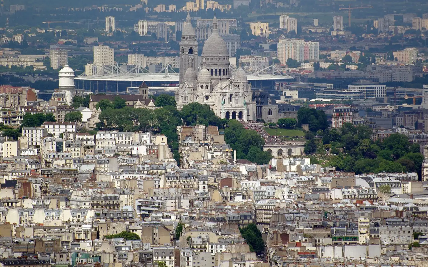 La basilique du Sacré-Cœur de Montmartre au premier plan, avec le toit du Stade de France qui se détache derrière elle au-dessus des immeubles