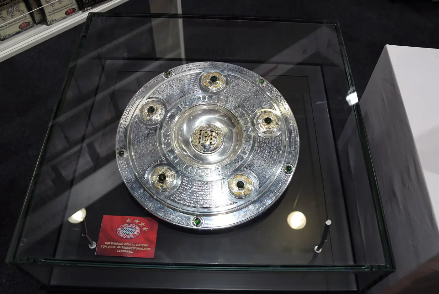 La Meisterschale, grand plat d'argent orné de pierres vertes et gravé des noms des champions, présentée sous une vitrine de verre avec un carton du Bayern Munich