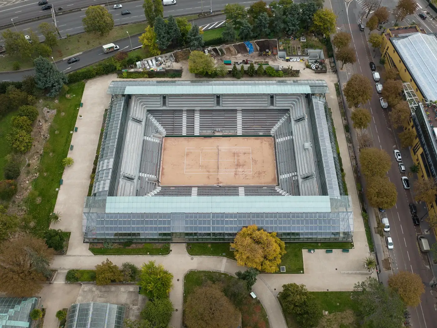 Le court Simonne-Mathieu vu du ciel : un court en terre battue entouré de tribunes et de serres vitrées, au milieu des arbres du jardin des serres d'Auteuil