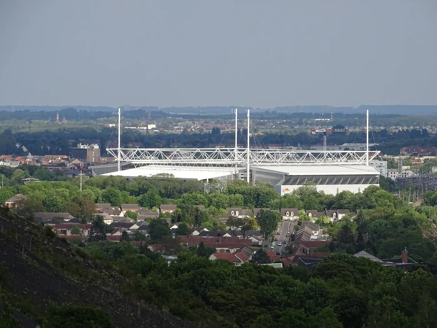 Le stade Bollaert-Delelis de Lens et sa charpente métallique blanche, vus de loin au-dessus des toits et des arbres de la ville