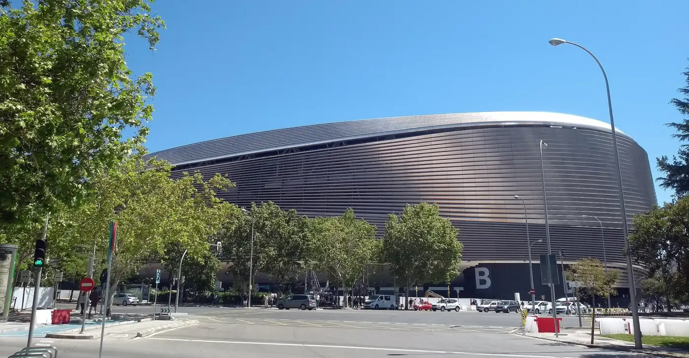Le stade Santiago-Bernabéu rénové, enveloppe métallique aux lames horizontales et toit courbe, vu depuis un carrefour bordé d'arbres à Madrid