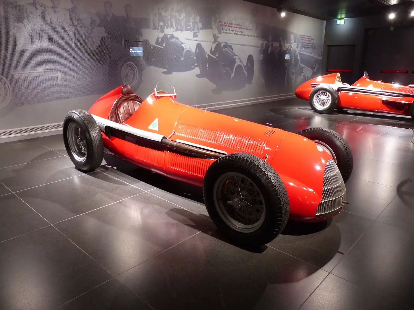 Monoplace rouge Alfa Romeo 158 de 1950 exposée dans une salle de musée au sol sombre, une autre Alfa Romeo de course visible à l'arrière-plan