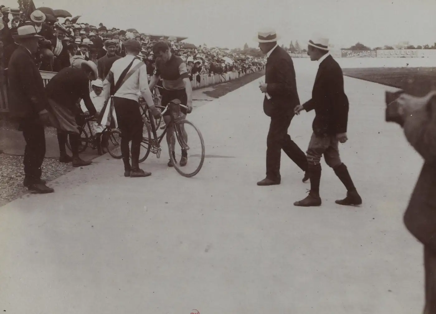 Photographie ancienne de l'arrivée du premier Tour de France sur une piste de vélodrome, un coureur tenant son vélo entouré d'officiels, tribunes pleines de spectateurs en canotier