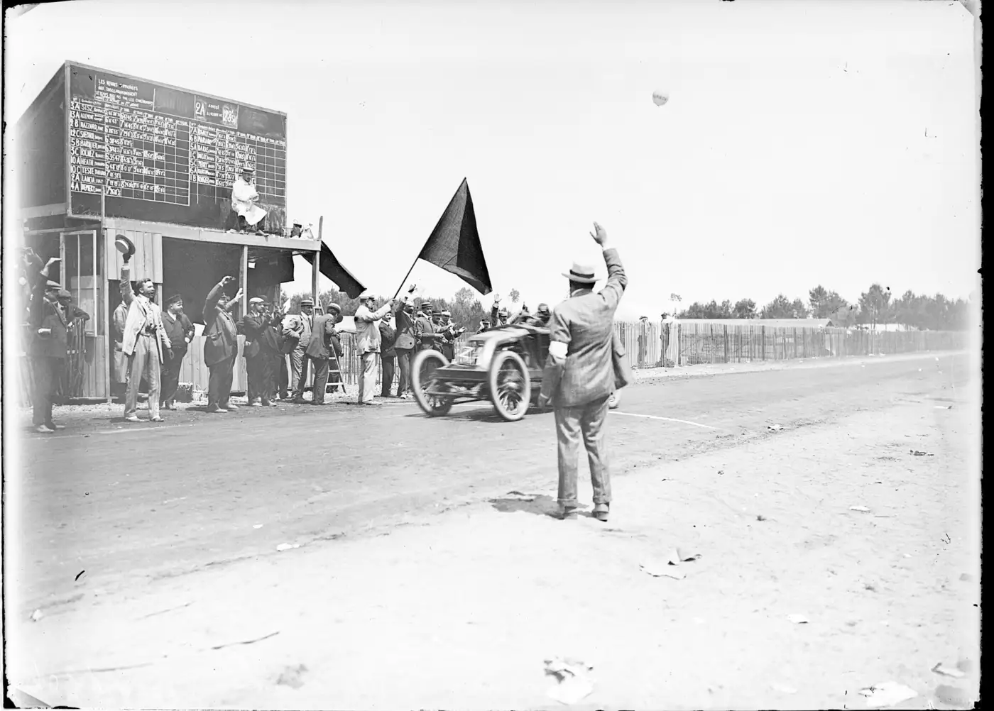 Photographie ancienne d'une voiture de course passant devant un panneau d'affichage des temps en bois, des officiels agitant chapeaux et drapeau au bord de la route