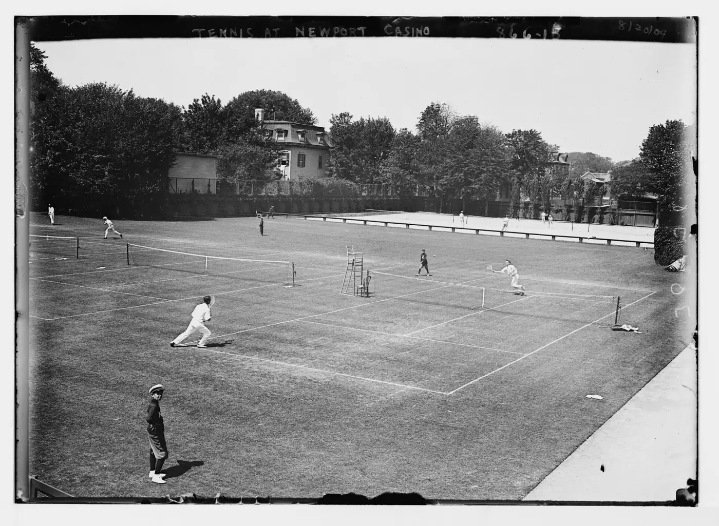 Photographie ancienne en noir et blanc des courts en gazon du Newport Casino, avec des joueurs en blanc, une chaise d'arbitre et un ramasseur de balles au premier plan