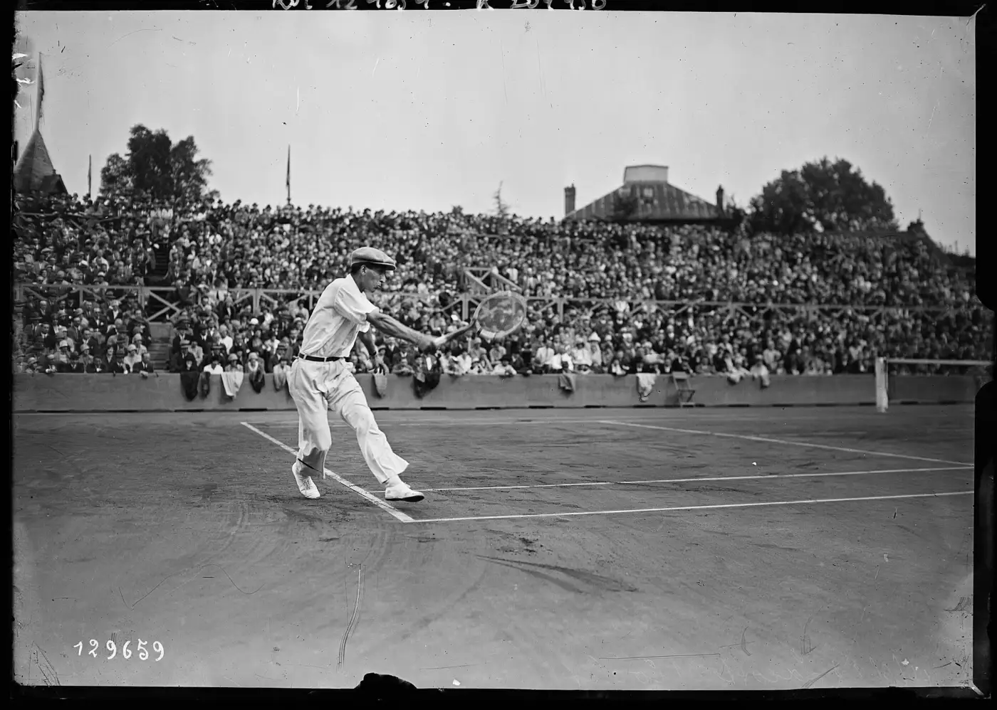 Photographie en noir et blanc de René Lacoste, en casquette et pantalon blanc, frappant un revers devant des tribunes combles à Roland-Garros en 1928