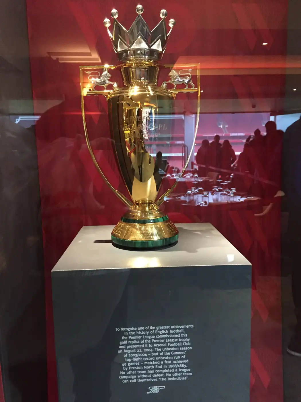 Réplique dorée du trophée de la Premier League, couronne et lions sur les anses, exposée sous vitrine sur un socle gris qui porte un texte sur la saison sans défaite d'Arsenal