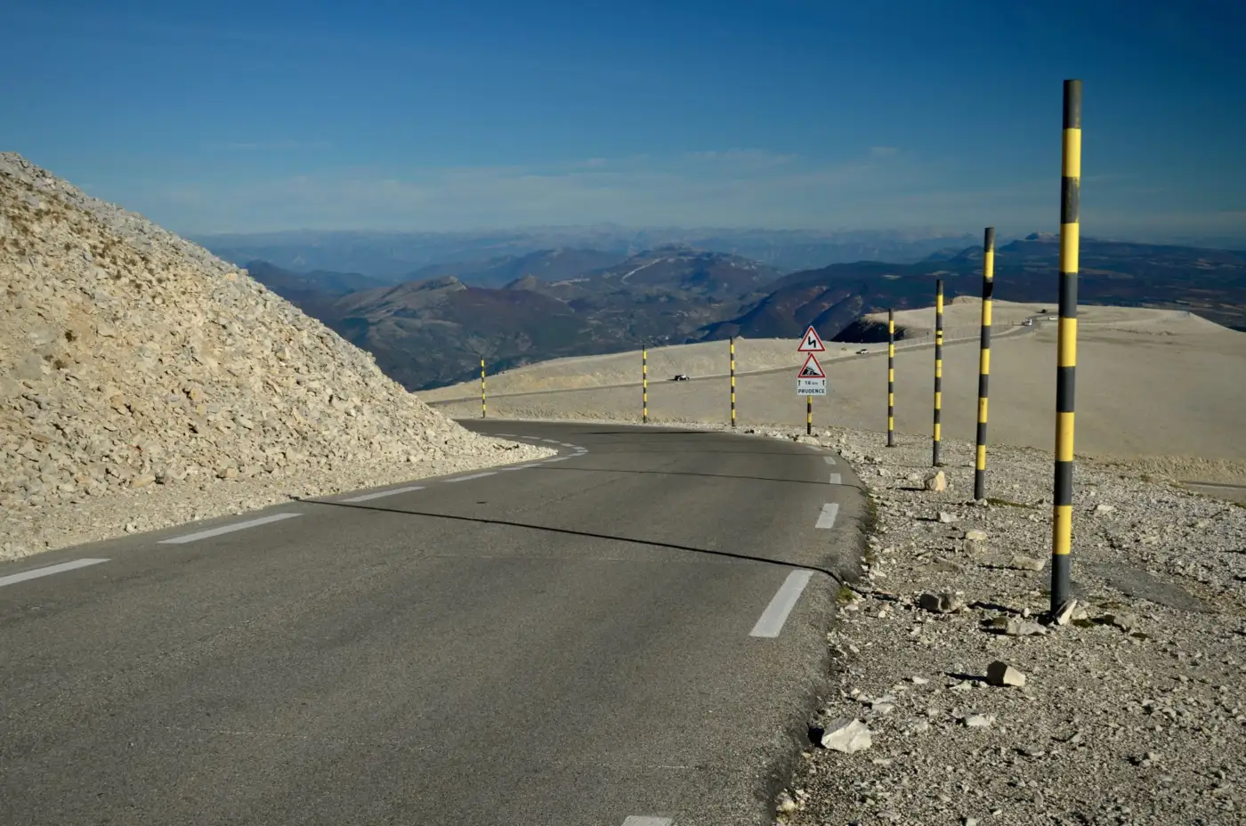 Route goudronnée en lacet près du sommet du mont Ventoux, bordée de pierres blanches et de piquets jaunes et noirs, montagnes de Provence à l'horizon