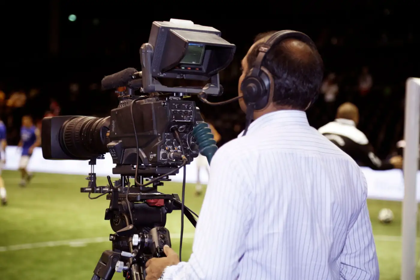 Un cadreur de télévision avec casque filme un match de football depuis le bord du terrain, de nuit
