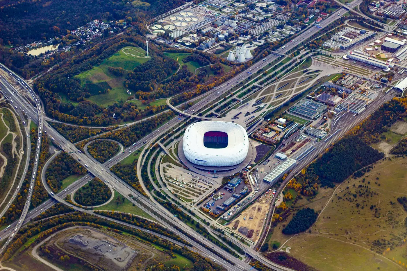 Vue aérienne de l'Allianz Arena de Munich, stade blanc aux coussins gonflés, entouré d'échangeurs autoroutiers et de bois aux couleurs d'automne