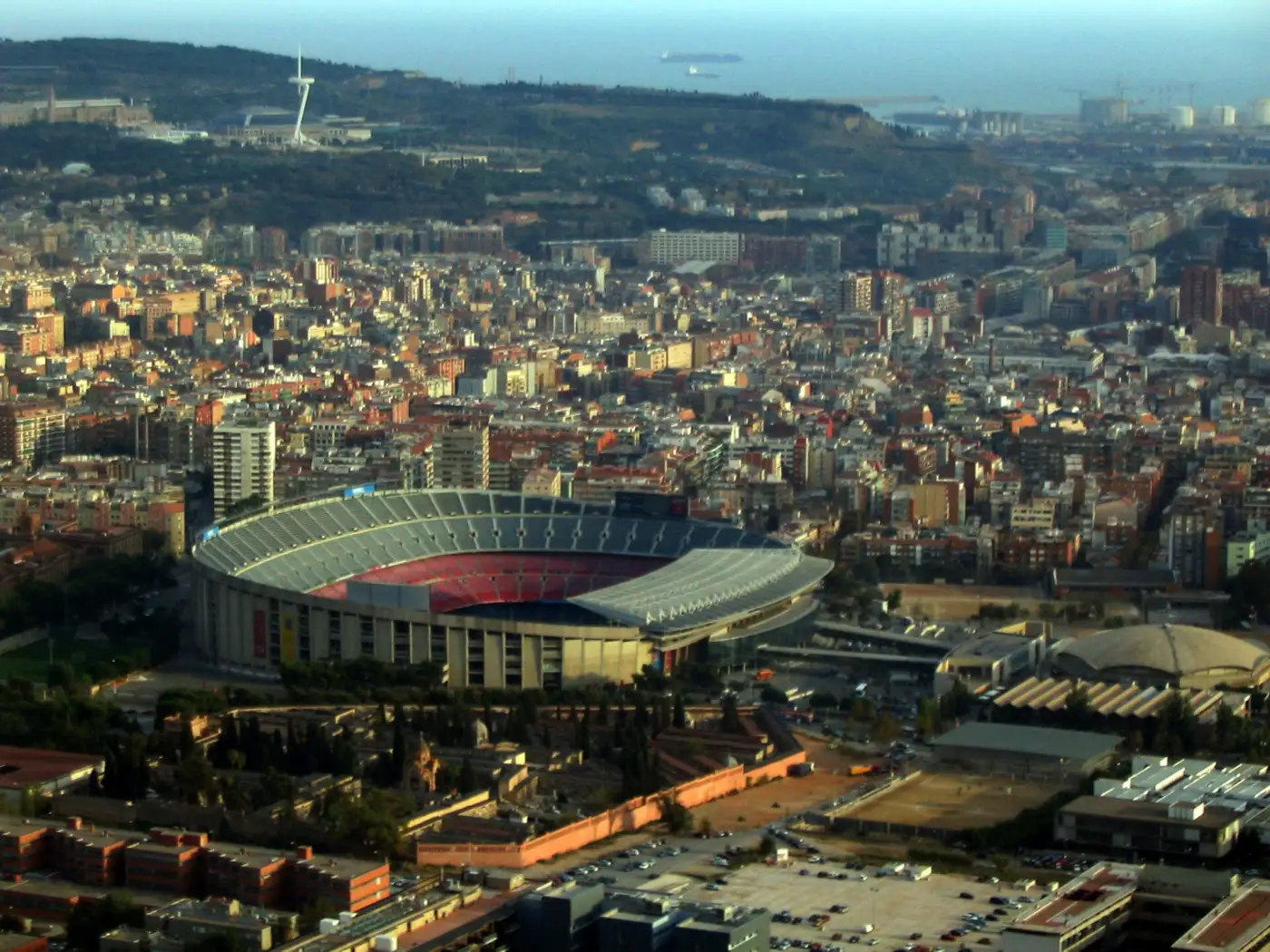 Vue aérienne du Camp Nou à Barcelone, cuvette aux sièges rouges et bleus au milieu des immeubles, la colline de Montjuïc et la mer en arrière-plan