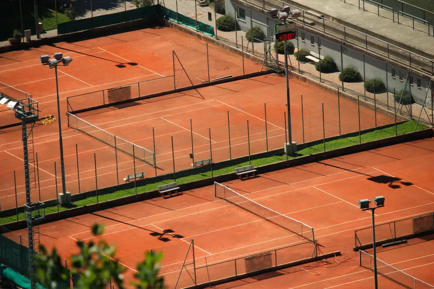 Vue plongeante sur des courts de tennis en terre battue vides, avec les ombres portées des filets et des projecteurs