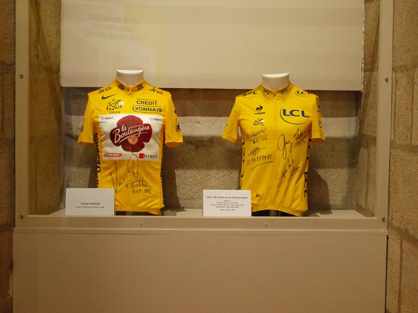 Deux maillots jaunes présentés sous vitrine dans une exposition, l'un de 2004 portant des logos de partenaires et une dédicace, l'autre de 2015 couvert de signatures