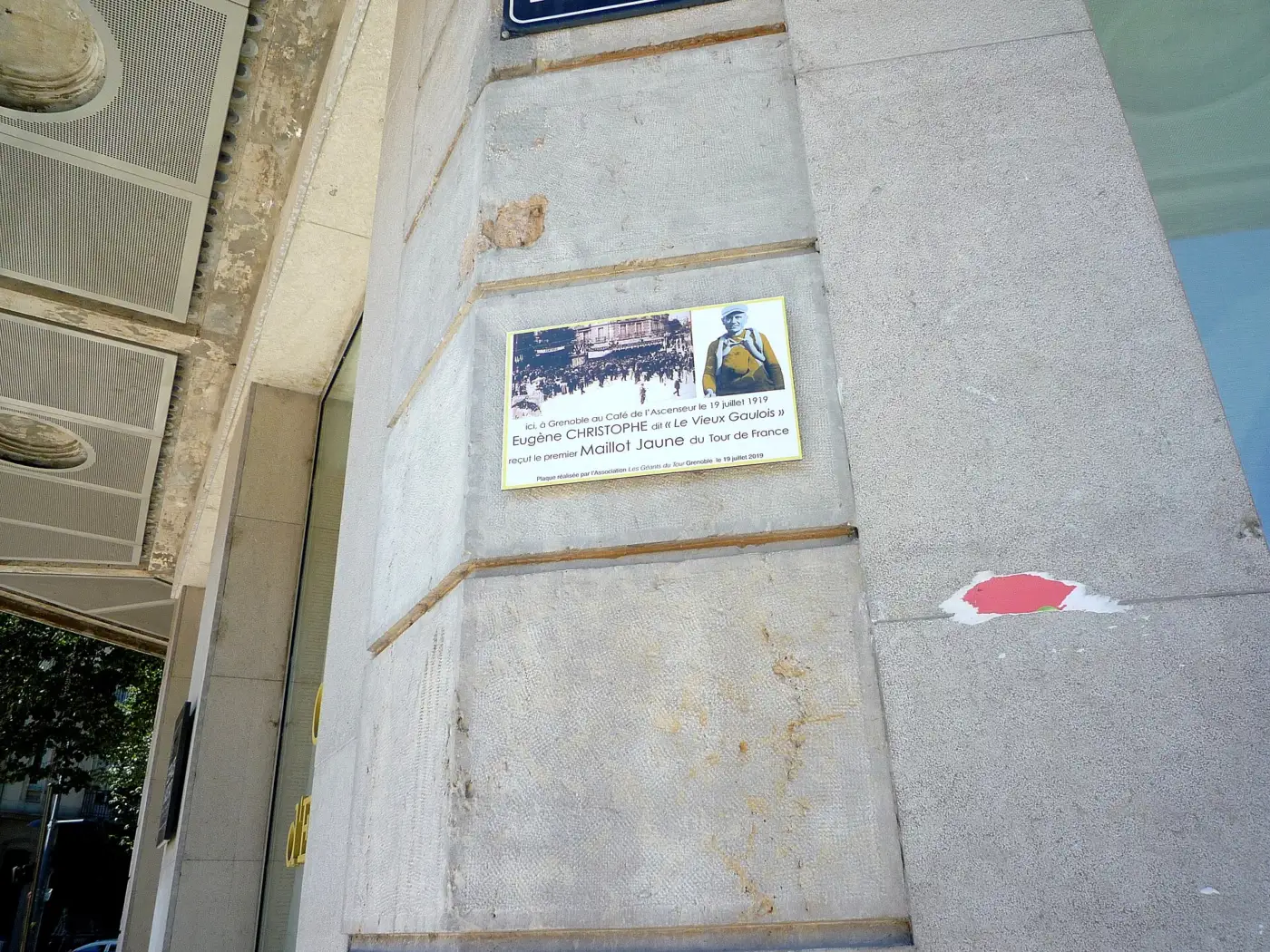 Plaque commémorative fixée sur un mur de pierre à Grenoble, avec une photo ancienne de foule et un portrait d'Eugène Christophe, rappelant la remise du premier maillot jaune en 1919