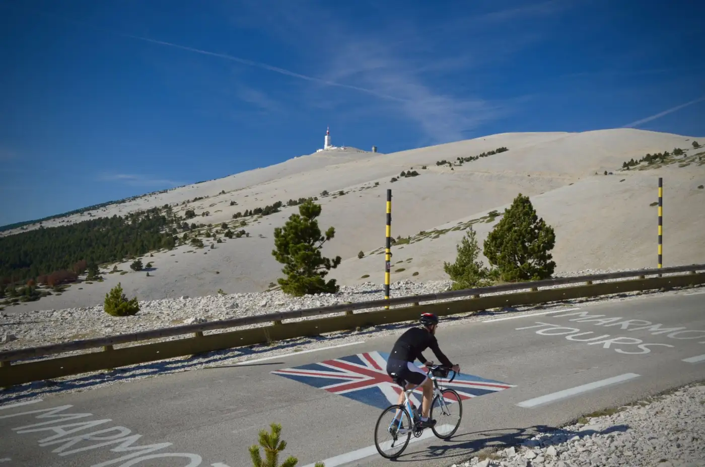 Un cycliste gravit la route du mont Ventoux, marquée de peintures au sol, avec le sommet pelé et son émetteur en arrière-plan sous un ciel bleu